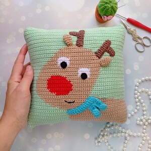 May include: A green crocheted pillow with a brown reindeer face on it. The reindeer has a red nose, white eyes, and a blue scarf.