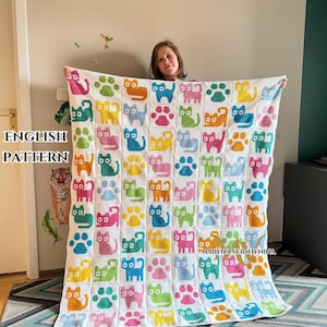 May include: A white crochet blanket featuring a repeating grid pattern of colorful cat silhouettes and paw prints in shades of pink, yellow, blue, green, and orange. The textile displays a consistent square block design throughout the entire piece.