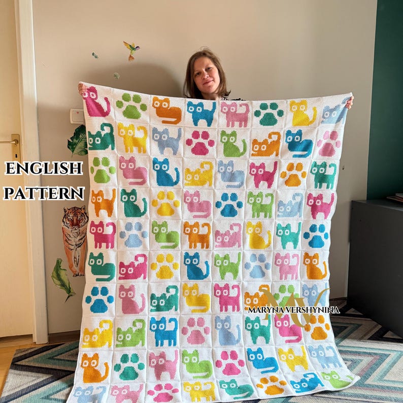 K&ouml;nnte beinhalten: Eine wei&szlig;e H&auml;keldecke mit einem sich wiederholenden Gittermuster aus bunten Katzensilhouetten und Pfotenabdr&uuml;cken in den Farben Pink, Gelb, Blau, Gr&uuml;n und Orange. Das Textil zeigt durchgehend ein einheitliches Design aus quadratischen Bl&ouml;cken.