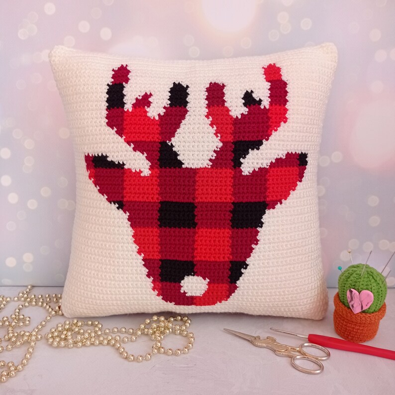 May include: A white crocheted pillow with a red and black buffalo plaid reindeer design. The pillow is sitting on a white surface with a string of gold beads and a small crocheted cactus with a pink heart.