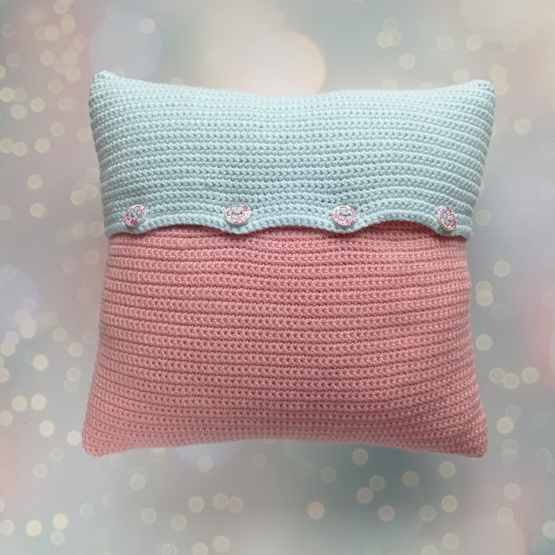 May include: A crocheted pillow with a light blue top and a pink bottom. The pillow has a scalloped edge with four buttons.