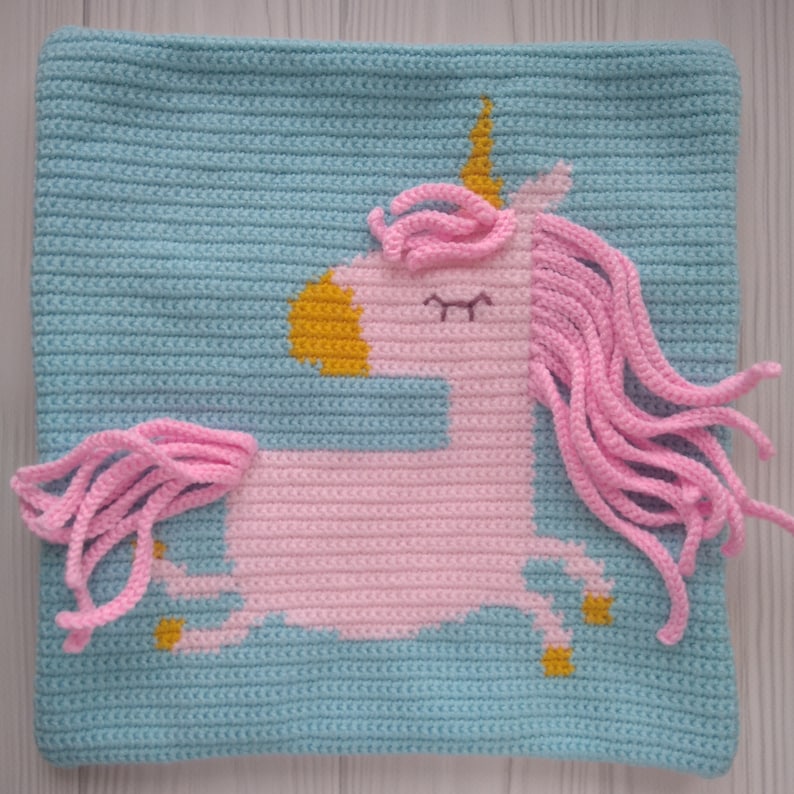 May include: A crocheted pillow with a pink unicorn design. The unicorn has a gold horn and a blue background.