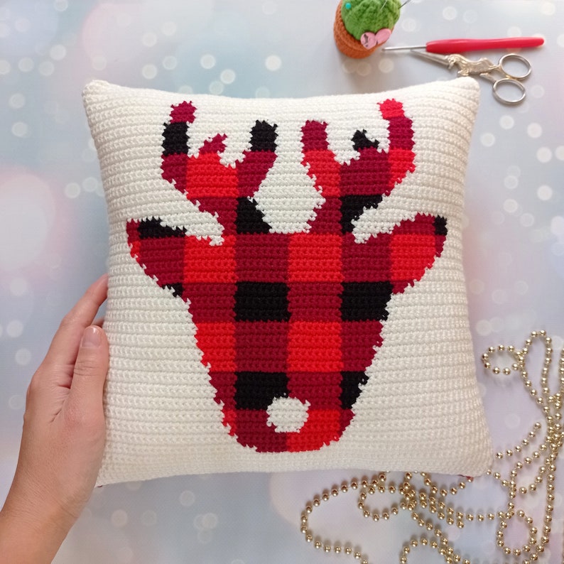 May include: A white crocheted pillow with a red and black buffalo plaid reindeer design.