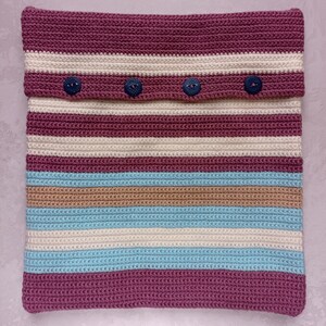 May include: A striped crocheted pillow with a burgundy, beige, white, and light blue color scheme. The pillow has four buttons on the top flap.