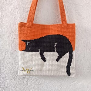 May include: A crocheted tote bag with a black cat design. The bag is orange and white with a black cat sleeping on the white portion. The bag has a black handle.