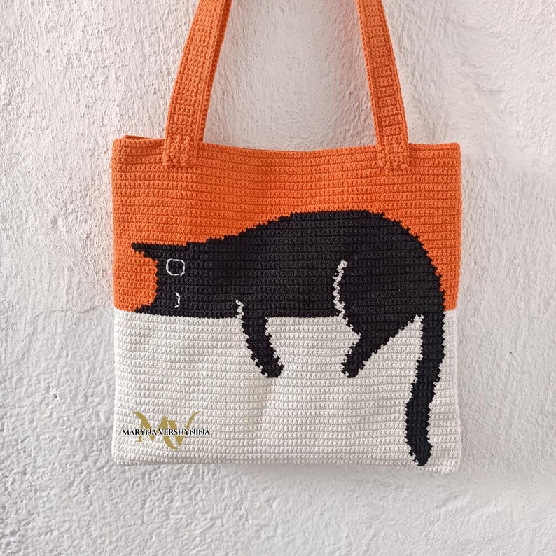 May include: A crocheted tote bag with a black cat design. The bag is orange and white with a black cat sleeping on the white portion. The bag has a black handle.