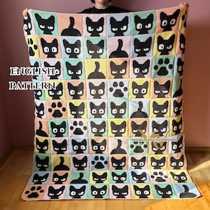 May include: A handmade crochet blanket featuring a repeating pattern of black cat faces and paw prints on a pastel color block background. The blanket has the text "ENGLISH PATTERN".