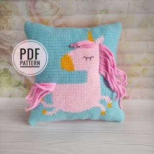 May include: A crochet pillow with a pink unicorn design. The unicorn has a yellow horn, yellow hooves, and pink hair. The pillow is on a light wood surface. PDF PATTERN.