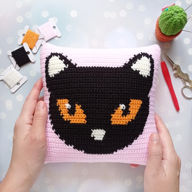May include: A pink crocheted pillow with a black cat face design. The cat has orange eyes and a white nose.