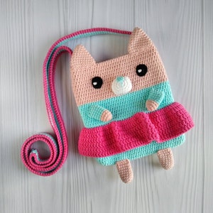 May include: A crocheted cat-shaped bag with a pink, turquoise, and white color scheme. The bag has a long strap and a ruffled skirt.