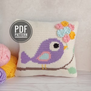 May include: A crocheted pillow featuring a purple bird with a pink and blue chest, perched on a brown branch with green leaves. The bird is holding a bouquet of pink, blue, and yellow flowers. The pillow is white with a "PDF PATTERN" label in the top left corner.