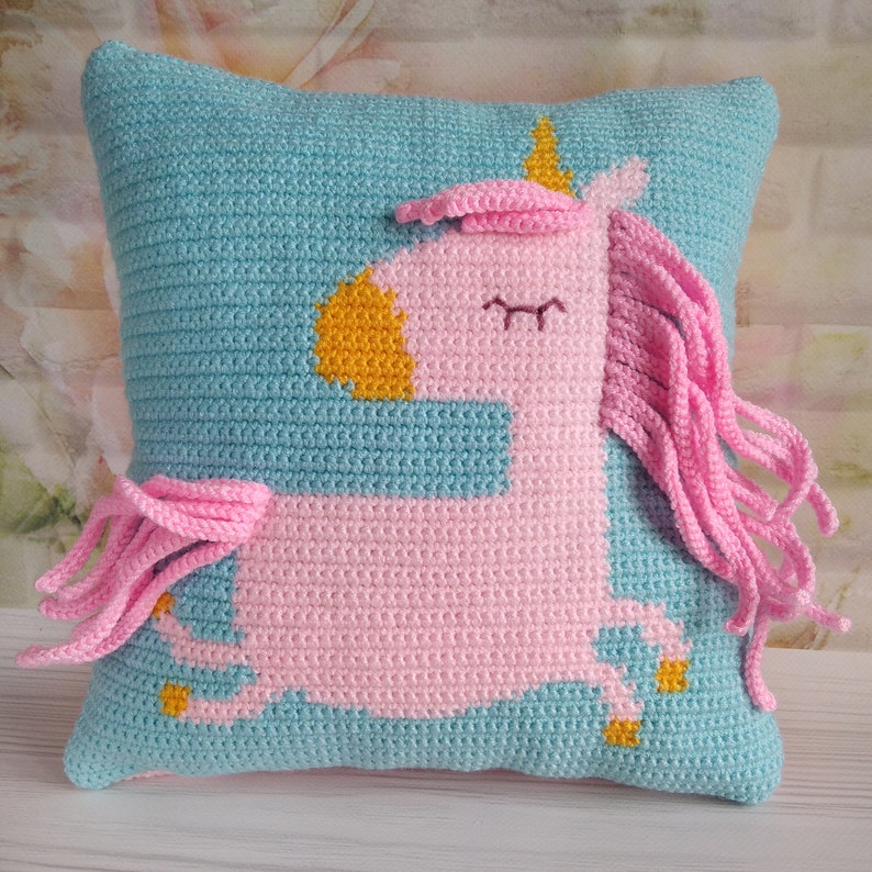 May include: A crocheted pillow with a pink unicorn design. The unicorn has a yellow horn, closed eyes, and a pink mane and tail.