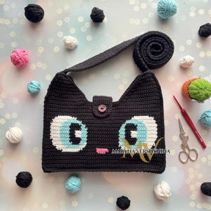 May include: A black crocheted cat-shaped handbag with blue eyes and a pink nose. The bag has a long strap and a button closure. The bag is surrounded by balls of yarn in various colors.