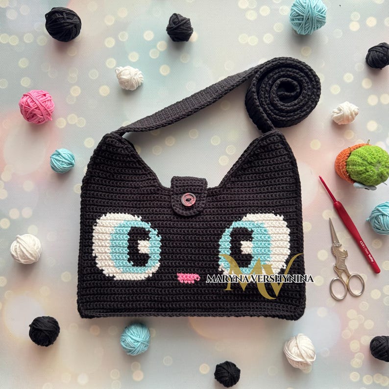 May include: A black crocheted cat-shaped handbag with blue eyes and a pink nose. The bag has a long strap and a button closure. The bag is surrounded by balls of yarn in various colors.