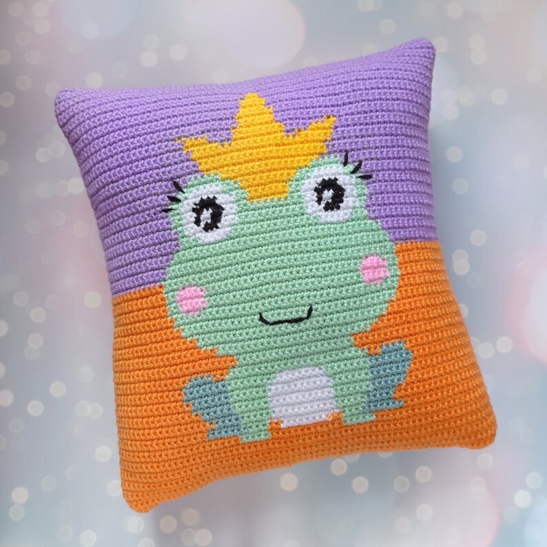 May include: A crocheted pillow with a green frog wearing a yellow crown. The frog has pink cheeks and black eyelashes. The pillow is purple on the top and orange on the bottom.