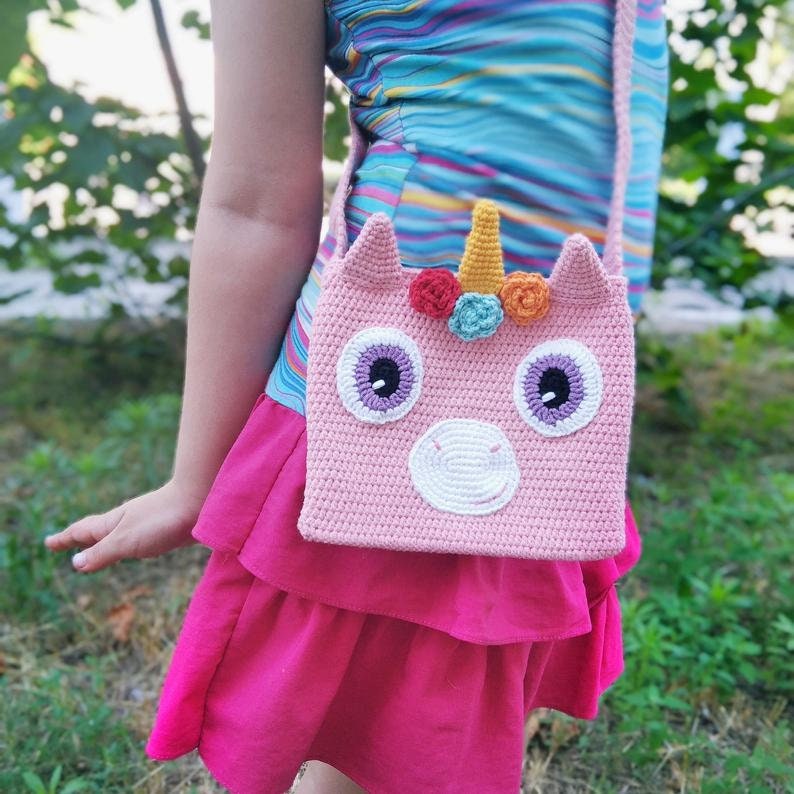 May include: A pink crocheted unicorn-shaped crossbody bag with a white horn, white muzzle, and purple eyes. The bag has colorful flowers on the top.