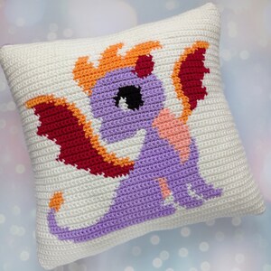 May include: A white crocheted pillow with a purple dragon design. The dragon has orange flames, red wings, and a pink belly.