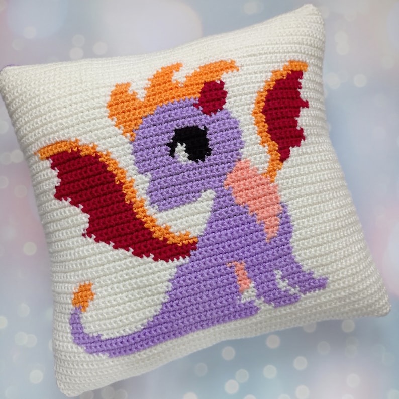 May include: A white crocheted pillow with a purple dragon design. The dragon has orange flames, red wings, and a pink belly.