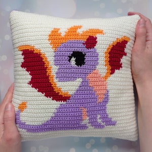 May include: A crocheted pillow with a purple dragon design. The dragon has orange flames, red wings, and a pink heart on its chest.