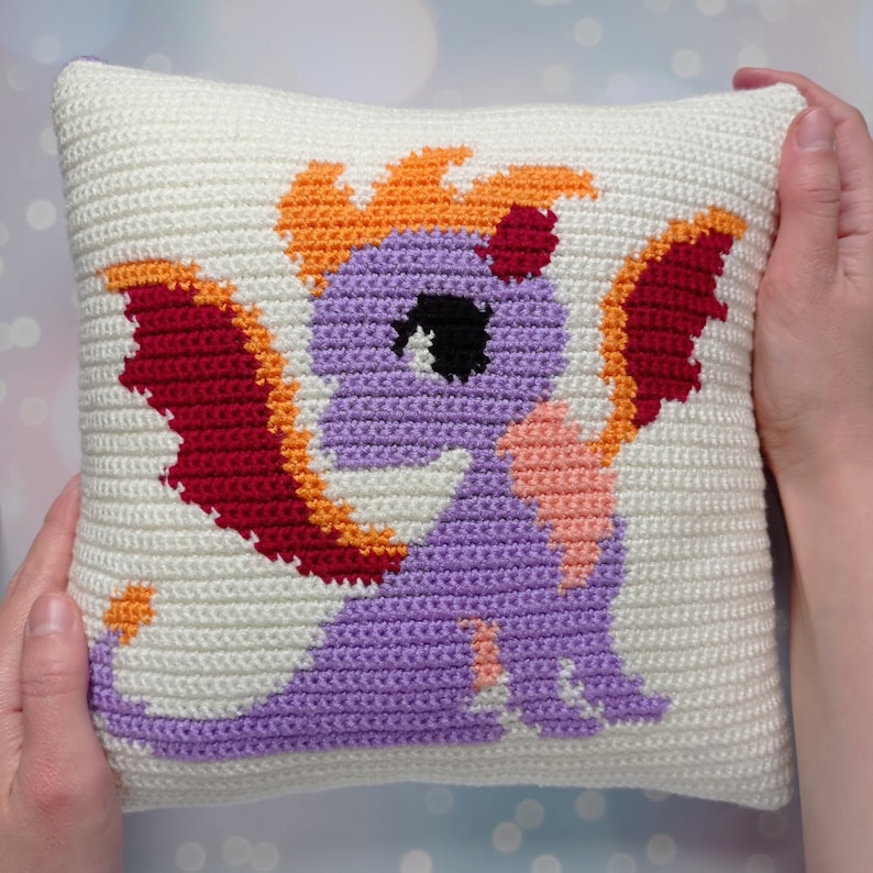 May include: A crocheted pillow with a purple dragon design. The dragon has orange flames, red wings, and a pink heart on its chest.
