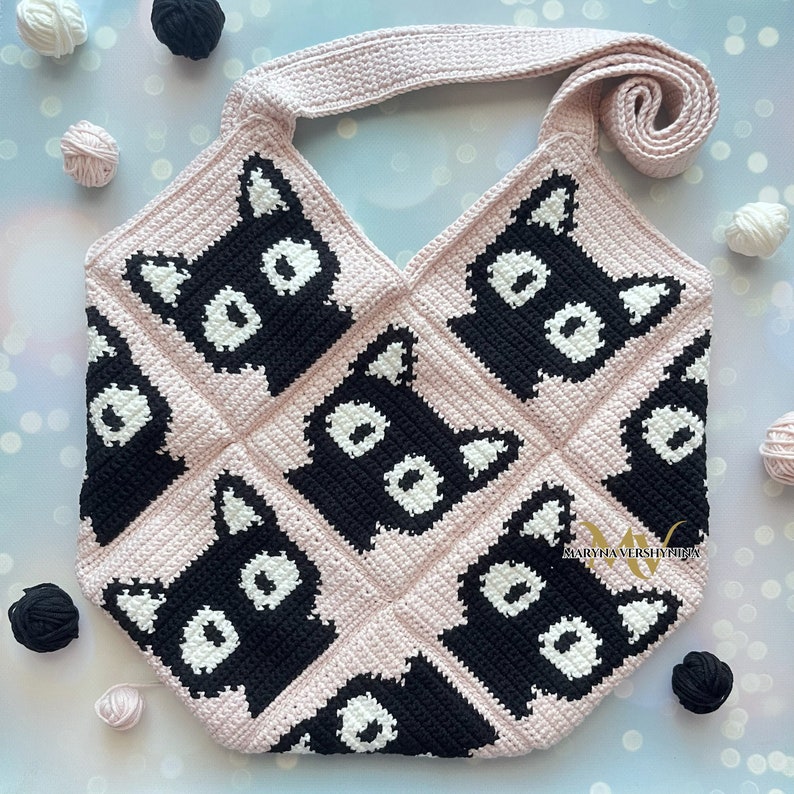 May include: A pink crocheted tote bag with black cat faces. The bag has a single strap and is made with a square stitch pattern.
