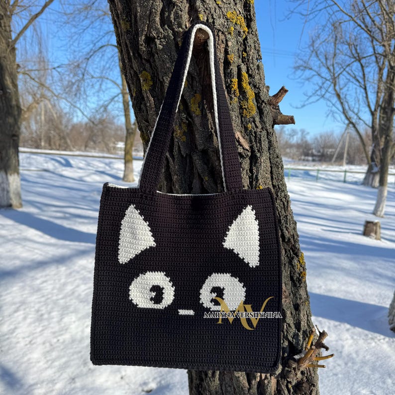 May include: Black crocheted tote bag with white crocheted cat face details. The cat face has two white ears, two white eyes, and a white nose.