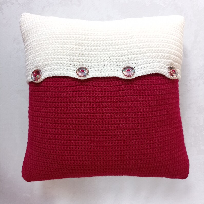 May include: A white and red crocheted pillow with a scalloped edge and four decorative buttons.