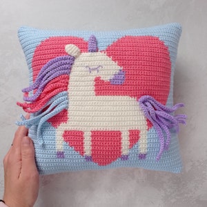 May include: A crocheted pillow with a pink heart and a white unicorn with purple and pink mane and tail.