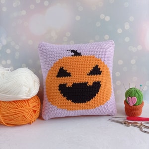 May include: A crocheted pillow with a purple background and an orange pumpkin with a black smiling face. The pillow is sitting on a white surface next to a small cactus and a pair of scissors.