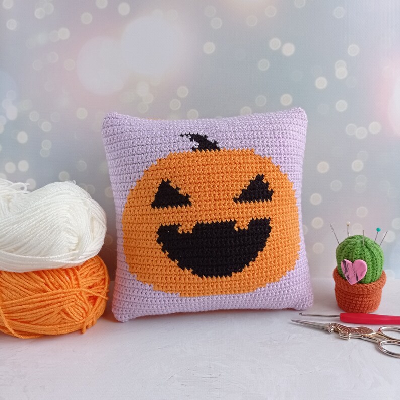May include: A crocheted pillow with a purple background and an orange pumpkin with a black smiling face. The pillow is sitting on a white surface next to a small cactus and a pair of scissors.