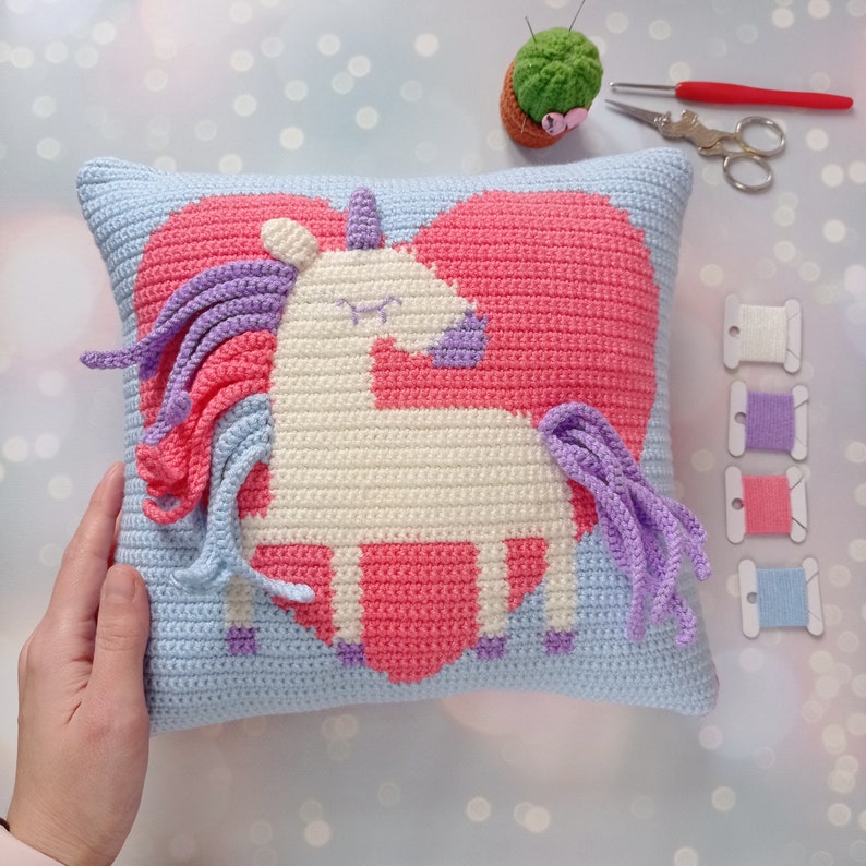 May include: A crocheted pillow with a light blue background and a pink heart with a white unicorn inside. The unicorn has a purple mane and tail.