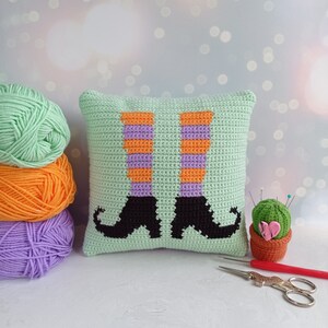 May include: A green crocheted pillow with a design of witch's legs in orange and purple stripes. The pillow is sitting on a white surface with yarn balls in the background.