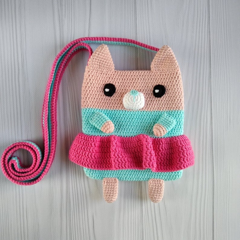 May include: A crocheted pink, teal, and white cat-shaped purse with a long strap. The purse has a pink skirt and black eyes.