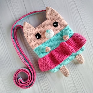 May include: A crocheted pink, blue, and white cat-shaped bag with a long strap. The bag has a pink and blue striped design and a white nose.