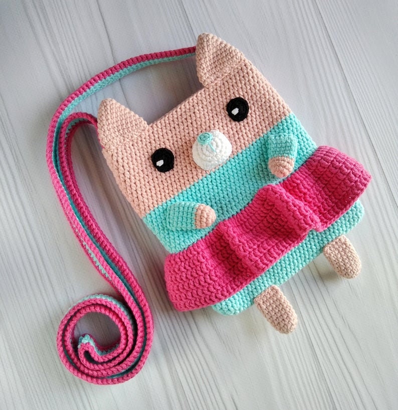 May include: A crocheted pink, blue, and white cat-shaped bag with a long strap. The bag has a pink and blue striped design and a white nose.