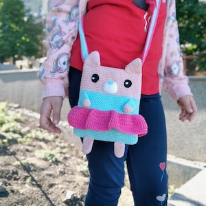 May include: A pink and blue crocheted purse shaped like a bear with a pink skirt. The purse has a strap and is being worn by a child.