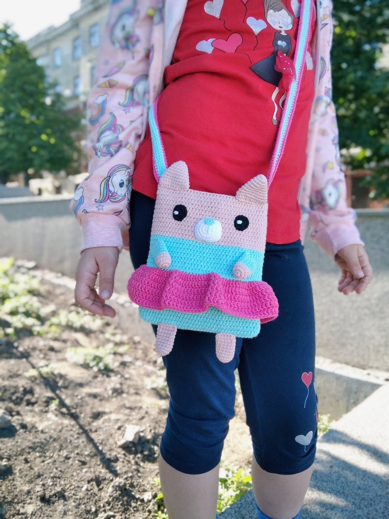 May include: A pink and blue crocheted purse shaped like a bear with a pink skirt. The purse has a strap and is being worn by a child.