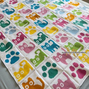 K&ouml;nnte beinhalten: Eine wei&szlig;e geh&auml;kelte Decke mit einem sich wiederholenden Gittermuster aus bunten Katzensilhouetten und Pfotenabdr&uuml;cken in Gelb, Rosa, Blau, Gr&uuml;n und Orange. Der Text MARYNA VERSHYNINA ist unten rechts in Schwarz auf die Decke gedruckt.