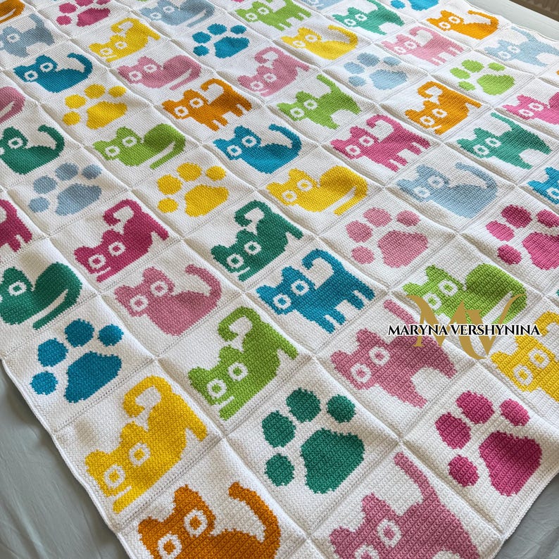 K&ouml;nnte beinhalten: Eine wei&szlig;e geh&auml;kelte Decke mit einem sich wiederholenden Gittermuster aus bunten Katzensilhouetten und Pfotenabdr&uuml;cken in Gelb, Rosa, Blau, Gr&uuml;n und Orange. Der Text MARYNA VERSHYNINA ist unten rechts in Schwarz auf die Decke gedruckt.