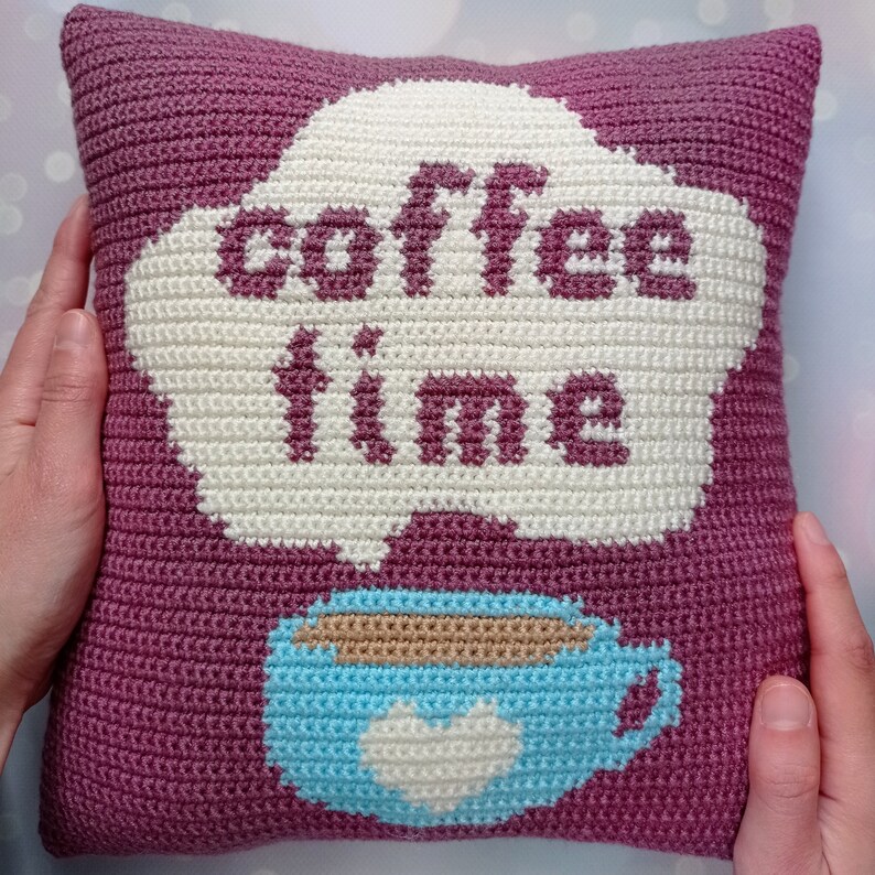 May include: A pink crocheted pillow with a white cloud-shaped applique that says "coffee time". A blue crocheted coffee cup with a white heart is below the cloud.