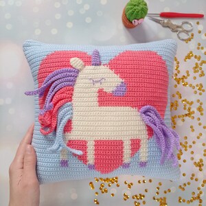 May include: A crocheted pillow with a light blue background and a pink heart with a white unicorn inside. The unicorn has a purple mane and tail.