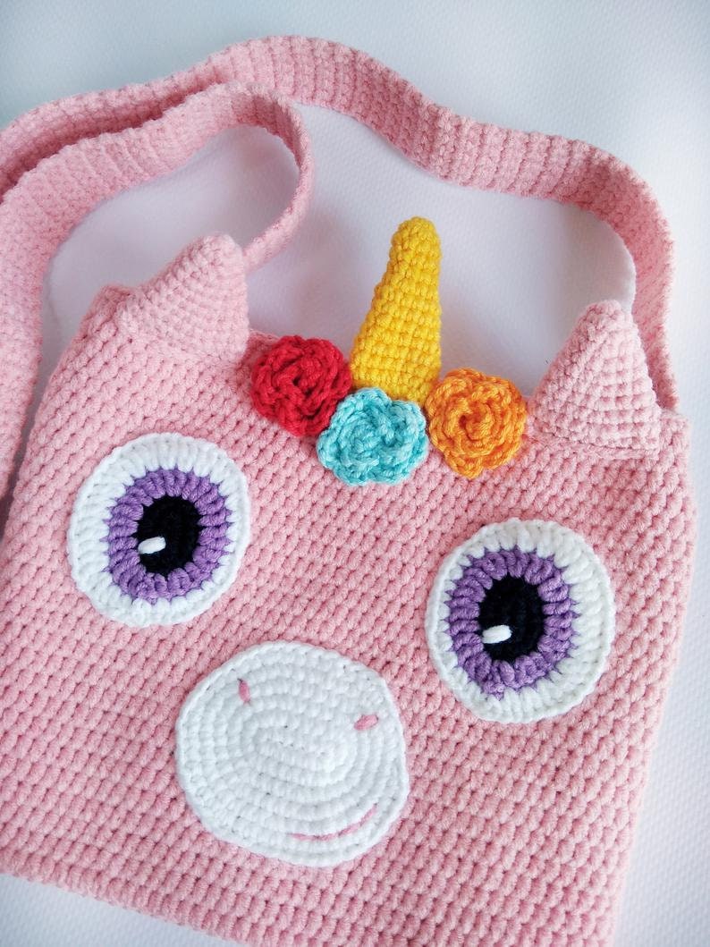 May include: A pink crocheted unicorn bag with a yellow horn, rainbow colored flowers, and purple and white eyes. The bag has a pink crocheted strap.