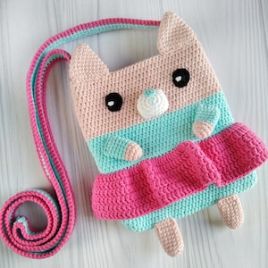 May include: A crocheted pink and turquoise cat-shaped bag with a long strap. The bag has a white nose and black eyes.