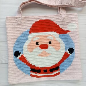 May include: A light pink crocheted tote bag with a Santa Claus design. The Santa Claus graphic is in red, white, black, and peach colours, set against a light blue circle. The bag has a matching pink handle.