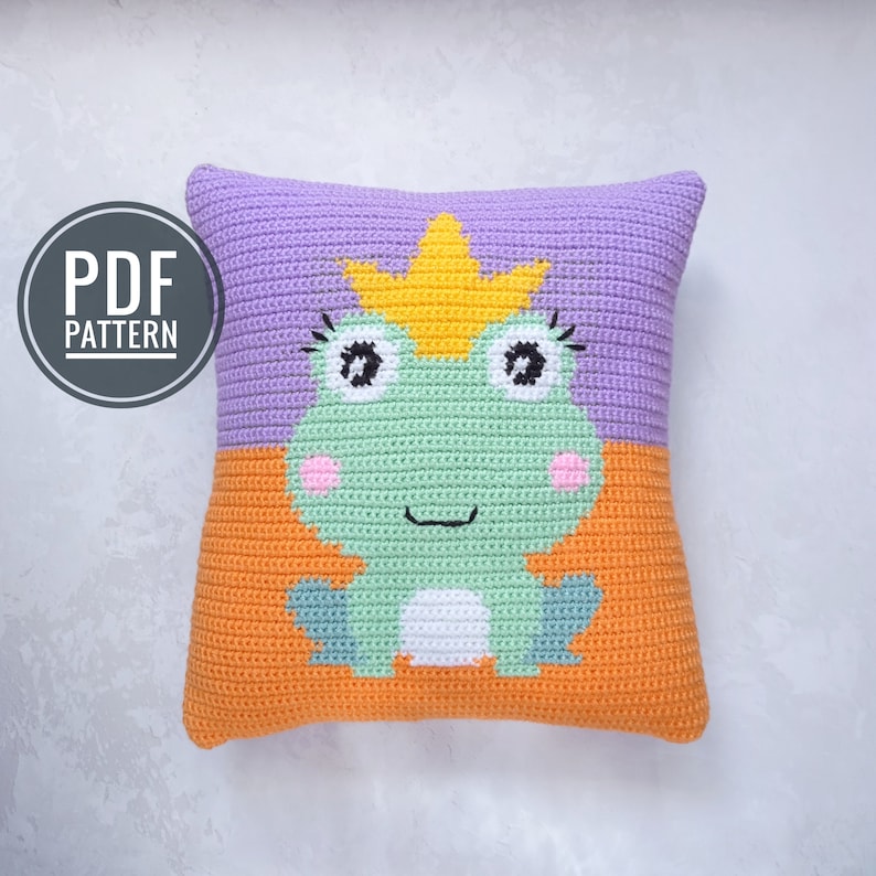 May include: Crochet pillow pattern featuring a green frog wearing a yellow crown. The pillow is made with purple and orange yarn.