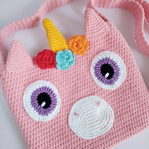 May include: A pink crocheted unicorn-shaped bag with a white crocheted nose, purple and white eyes, and a yellow horn with rainbow colored flowers. The bag has a long pink crocheted strap.