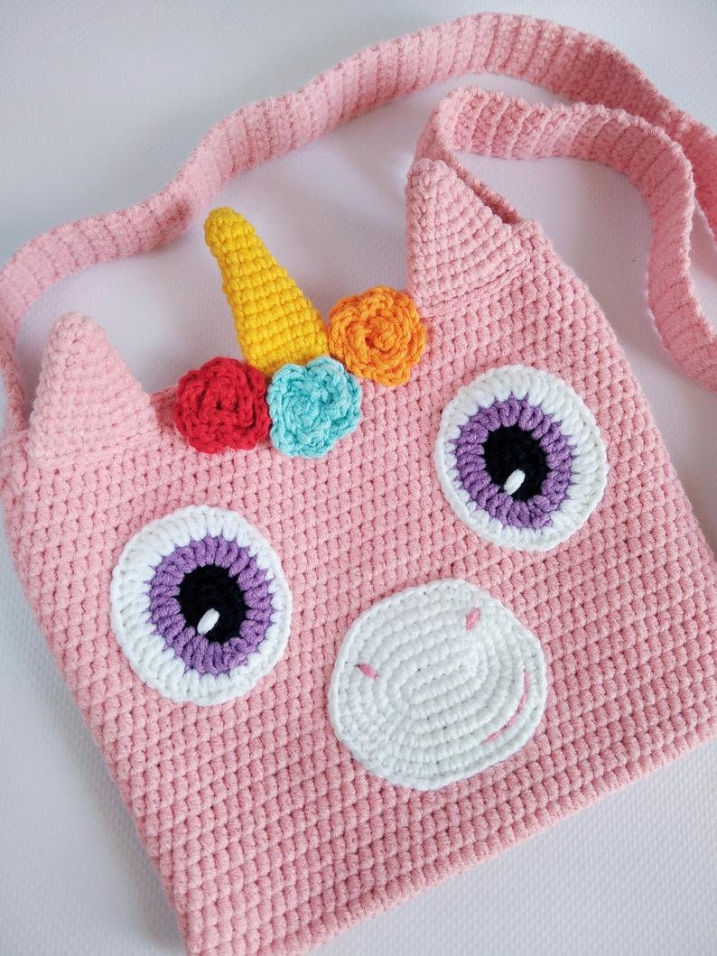 May include: A pink crocheted unicorn-shaped bag with a white crocheted nose, purple and white eyes, and a yellow horn with rainbow colored flowers. The bag has a long pink crocheted strap.