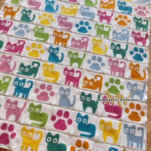 K&ouml;nnte beinhalten: Eine wei&szlig;e H&auml;keldecke mit einem sich wiederholenden Gittermuster aus bunten Katzen und Pfotenabdr&uuml;cken in den Farben Pink, Gelb, Blau, Gr&uuml;n und Orange. Der Text MARYNA VERSHYNINA ist unten rechts auf der Decke in Schwarz aufgedruckt.