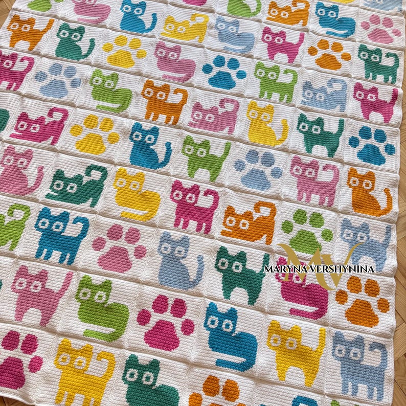 K&ouml;nnte beinhalten: Eine wei&szlig;e H&auml;keldecke mit einem sich wiederholenden Gittermuster aus bunten Katzen und Pfotenabdr&uuml;cken in den Farben Pink, Gelb, Blau, Gr&uuml;n und Orange. Der Text MARYNA VERSHYNINA ist unten rechts auf der Decke in Schwarz aufgedruckt.