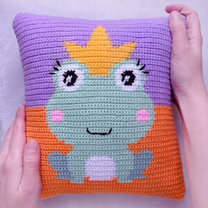 May include: A crocheted pillow with a green frog wearing a yellow crown. The frog has black eyes, a pink nose, and a white belly. The pillow is made of purple, orange, and green yarn.